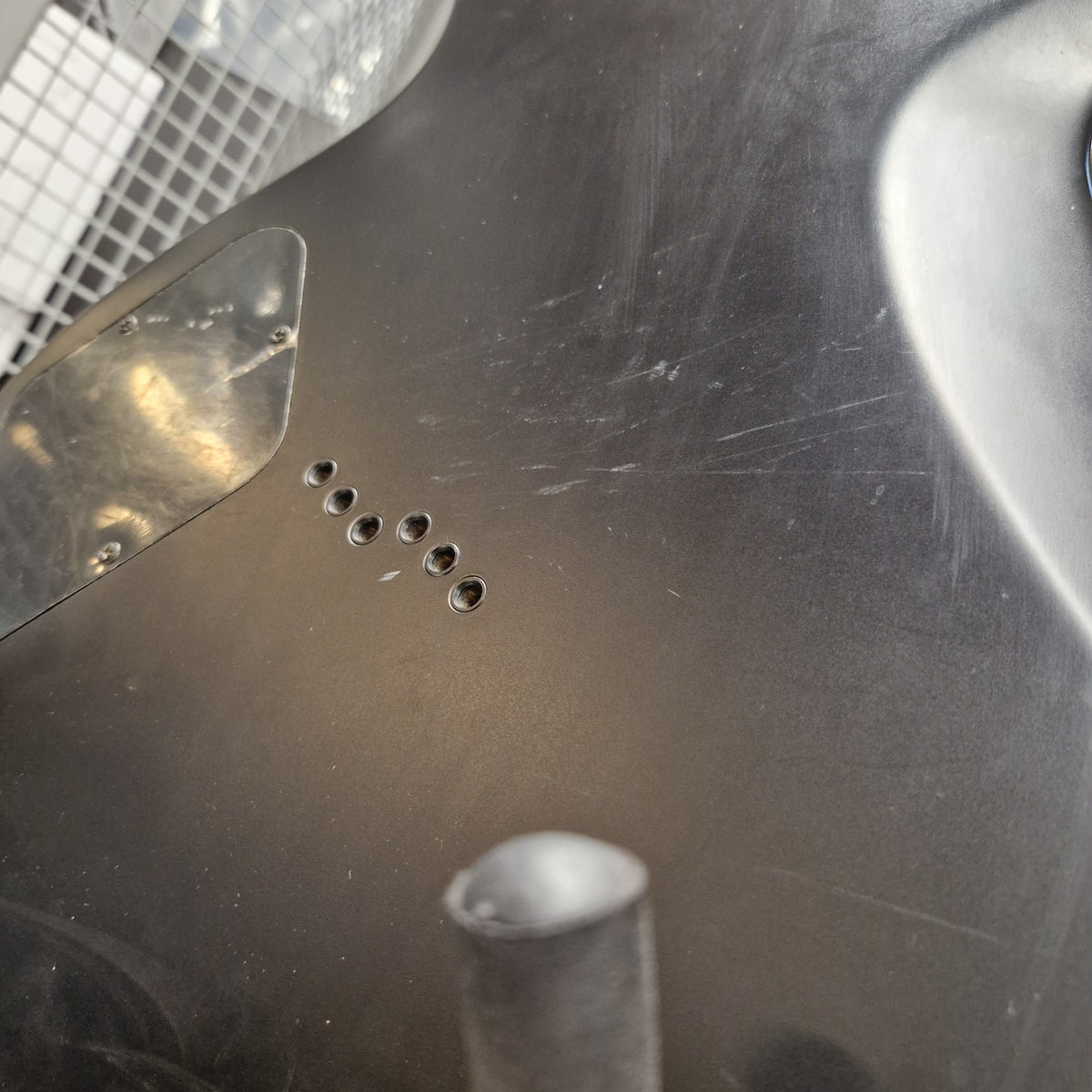 Pre-Owned Cort EVL-Z4 Electric - Satin Black Close-up of a dark metallic surface with a series of five small circular holes and a reflective brushed metal texture.