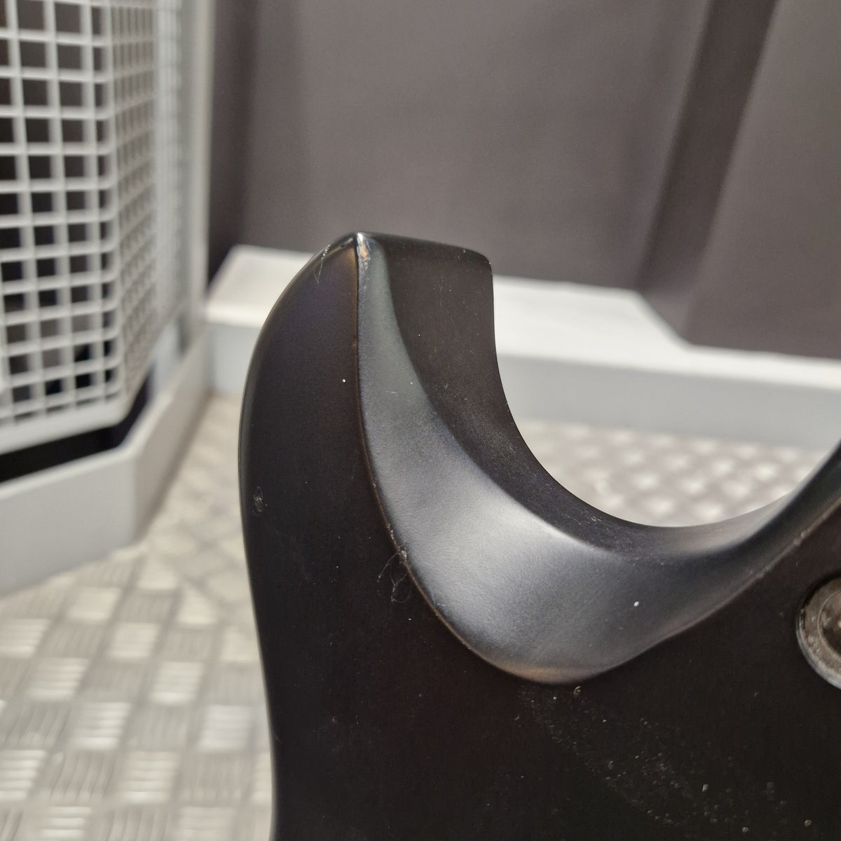 Pre-Owned Cort EVL-Z4 Electric - Satin Black Close-up of a black curved plastic component, likely a handle or bracket, showing a worn or scratched edge against a gray and white industrial background.