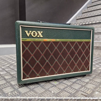 Pre-Owned Vox Pathfinder 10 Electric Amplifier Combo A vintage-style Vox guitar amplifier cabinet with a dark green textured finish, a brown diamond-pattern speaker grille, and gold 'VOX' lettering, photographed on a diamond-plate metal surface. 