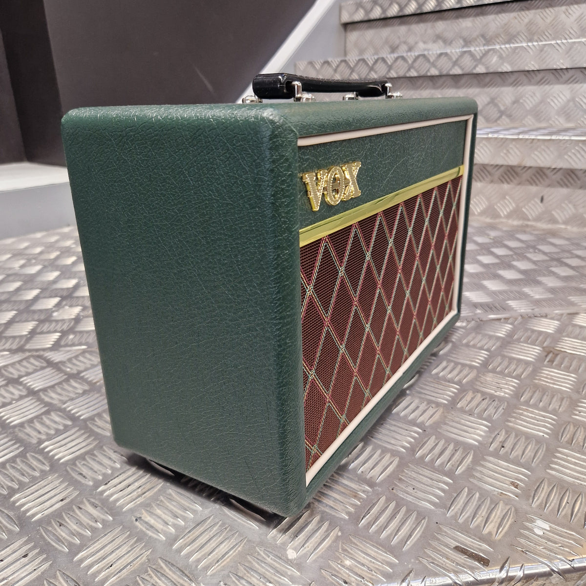 Pre-Owned Vox Pathfinder 10 Electric Amplifier Combo A green guitar amplifier head with a gold and red diamond-patterned grille and a gold 'Gibson' logo, photographed against a textured silver metal wall.