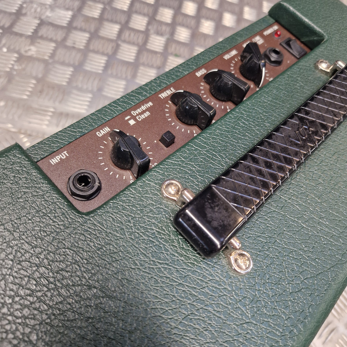 Pre-Owned Vox Pathfinder 10 Electric Amplifier Combo Close-up of a green textured amplifier head with black control knobs, a black handle, and a brown control panel, photographed against a metal diamond-plate floor.