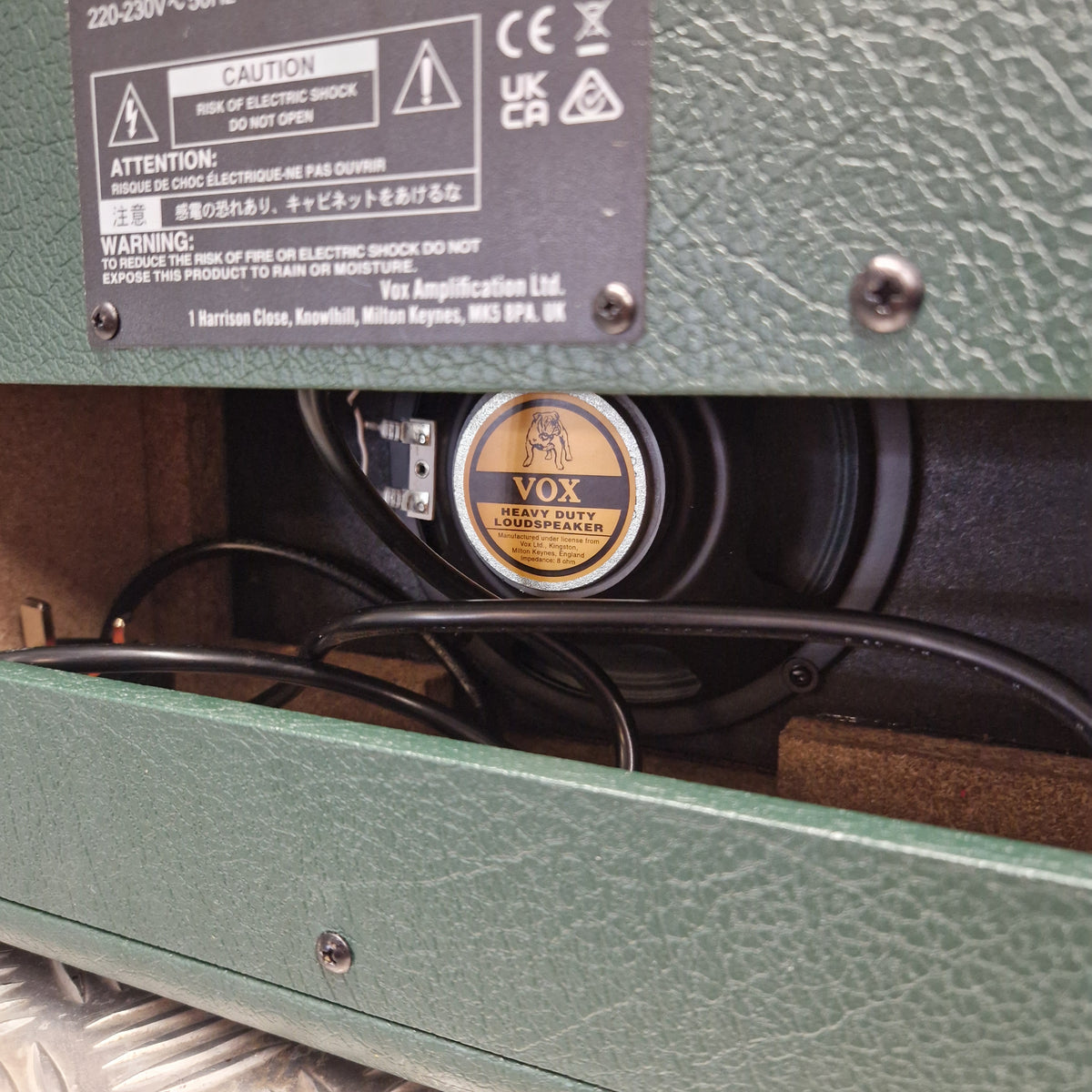Pre-Owned Vox Pathfinder 10 Electric Amplifier Combo Close-up of a green textured speaker cabinet showing a round black and gold VOX speaker inside, with black cables and a black specification label on the left.