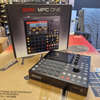 Pre-Owned Akai MPC ONE Standalone Music Production Centre AKAI MPC One digital music production device shown next to its retail box, photographed in a store or warehouse setting with cardboard boxes and other equipment in the background.