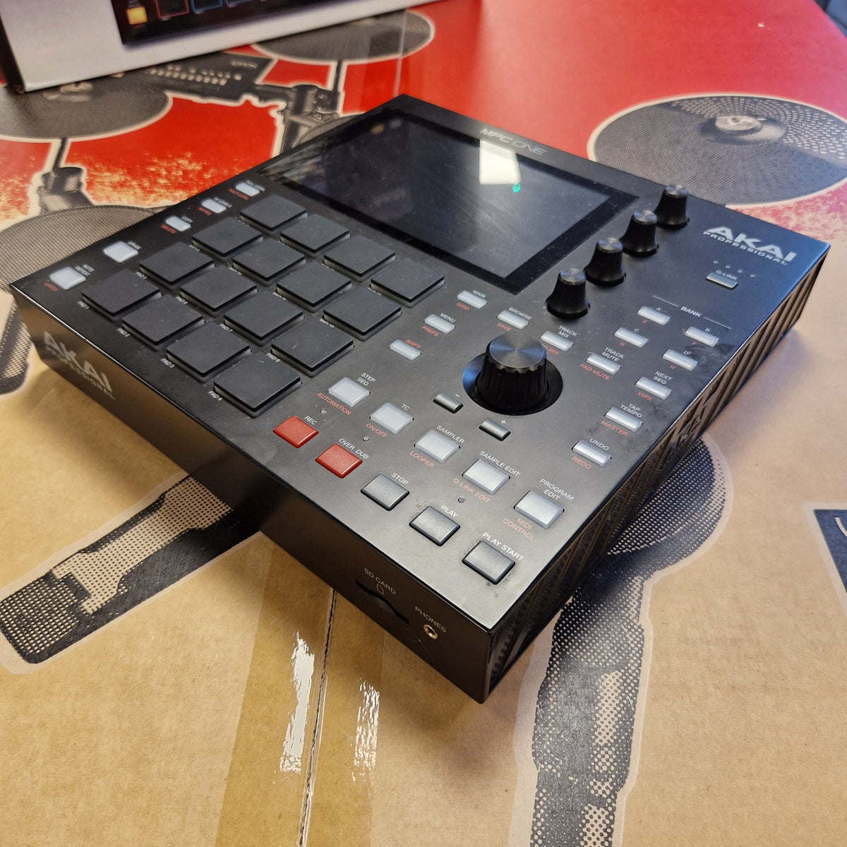 Pre-Owned Akai MPC ONE Standalone Music Production Centre An Akai DNX1000 portable music production controller in black, photographed at an angle on top of its cardboard retail packaging.