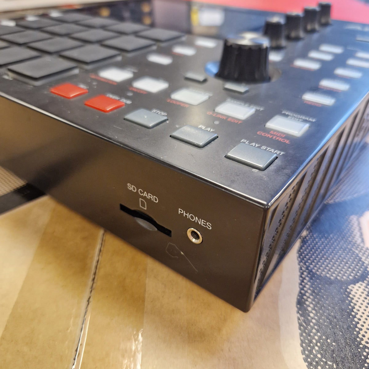 Pre-Owned Akai MPC ONE Standalone Music Production Centre A close-up angled view of a black electronic music controller or mixer, showing buttons, rotary knobs, and ports including a headphone jack and 3.5mm card slot, resting on a cardboard surface.