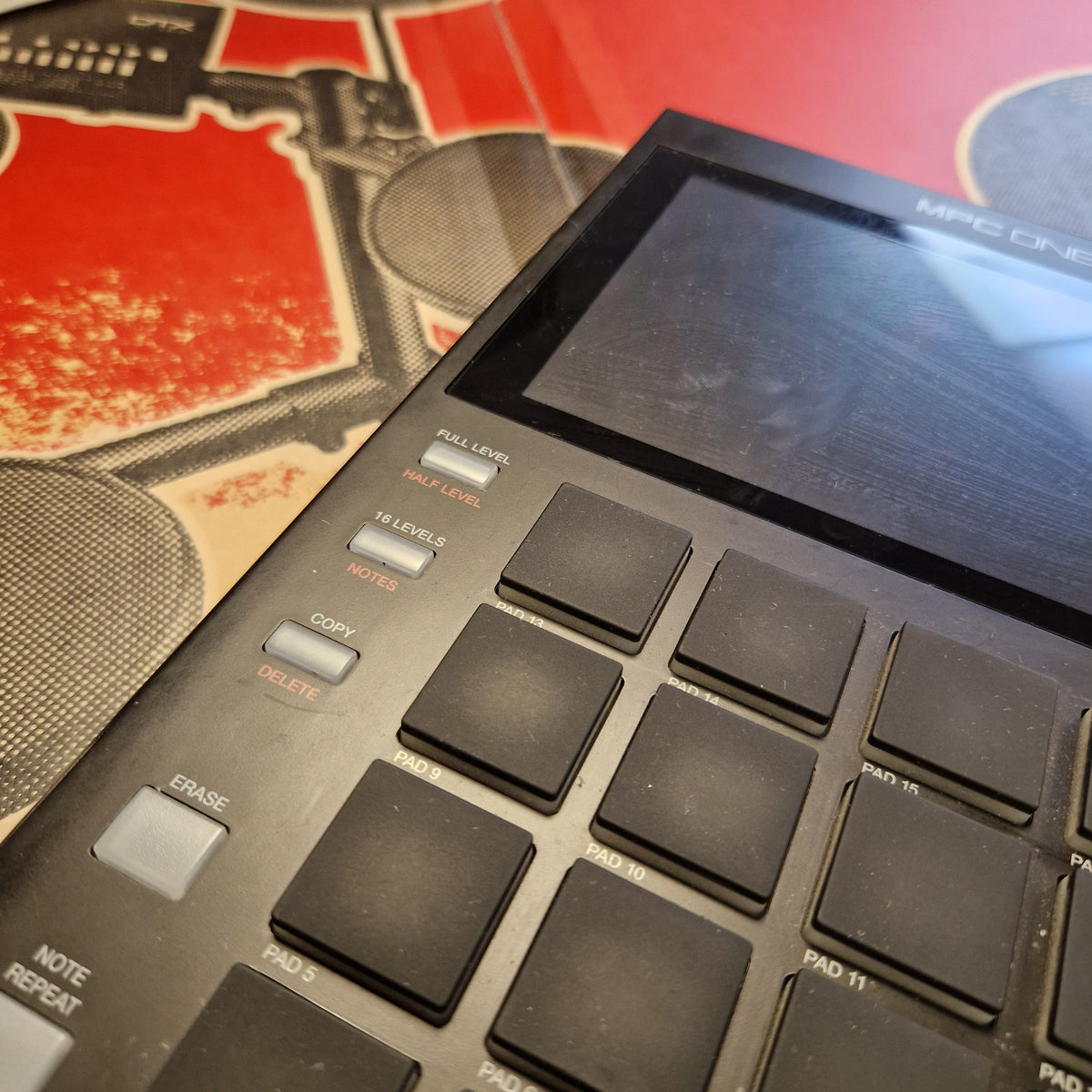 Pre-Owned Akai MPC ONE Standalone Music Production Centre A close-up angled view of a black MPC electronic music production controller with a screen and labeled buttons, resting on a red and black printed surface.