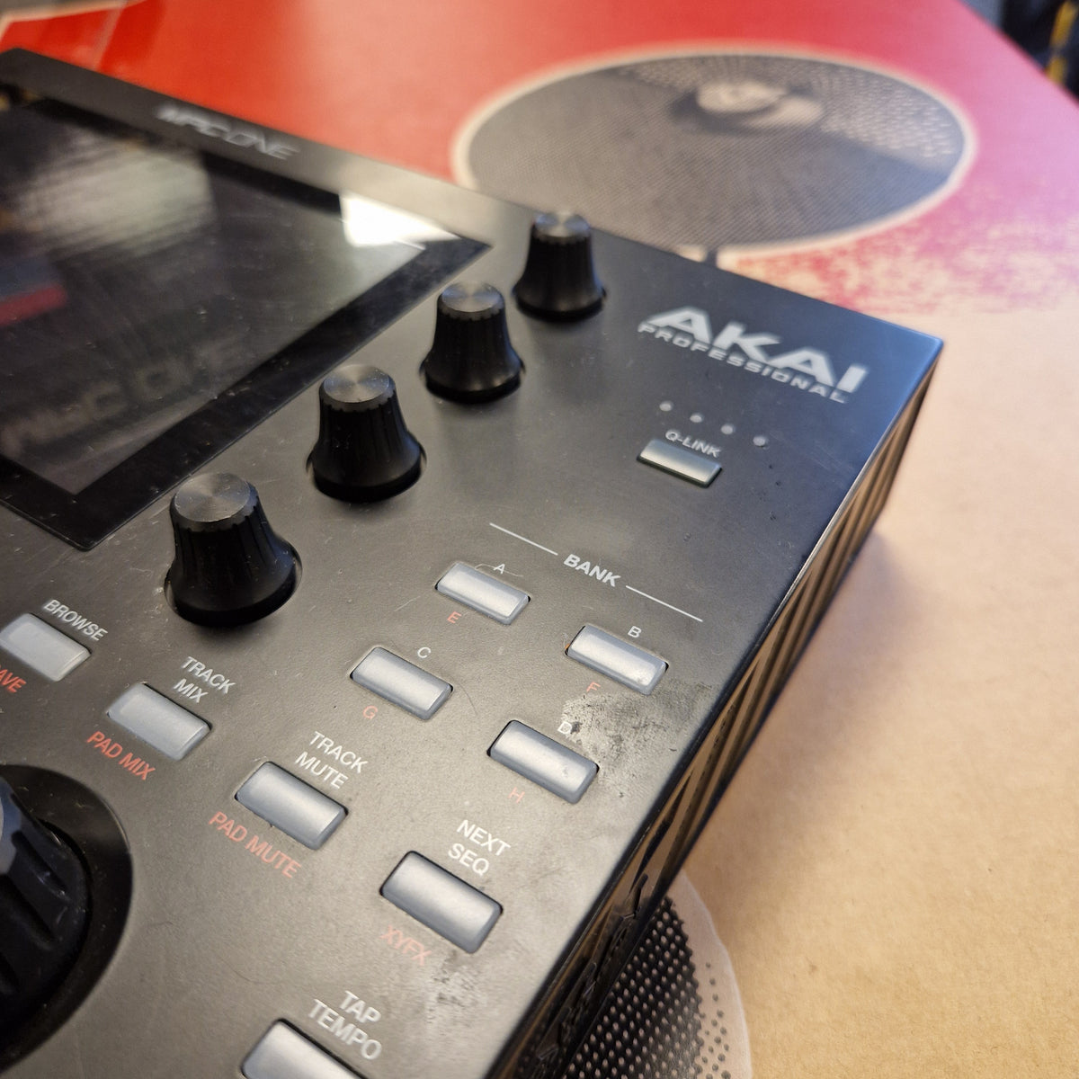 Pre-Owned Akai MPC ONE Standalone Music Production Centre Close-up of an Akai Professional audio controller or mixer unit with black knobs, silver buttons, and a display screen, photographed on a beige surface with part of a red speaker visible in the background.