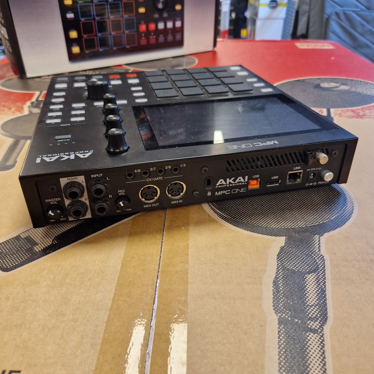 Pre-Owned Akai MPC ONE Standalone Music Production Centre An AAKI Uplouds 8-step sequencer and DAW controller device, shown at an angle on top of its original cardboard packaging box.