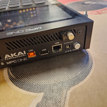 Pre-Owned Akai MPC ONE Standalone Music Production Centre Close-up of the rear panel of an Akai MPC One professional mixing device, showing ports, knobs, and branding, resting on a patterned surface.