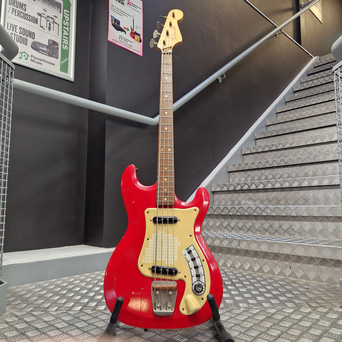 Pre-Owned Hagstrom Futurama Deluxe 1960s Bass - Red A red electric bass guitar with a cream pickguard and headstock, mounted on a display stand in a metal staircase area.