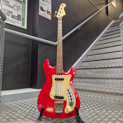 Pre-Owned Hagstrom Futurama Deluxe 1960s Bass - Red A red electric bass guitar with a cream pickguard and headstock, mounted on a display stand in a metal staircase area.