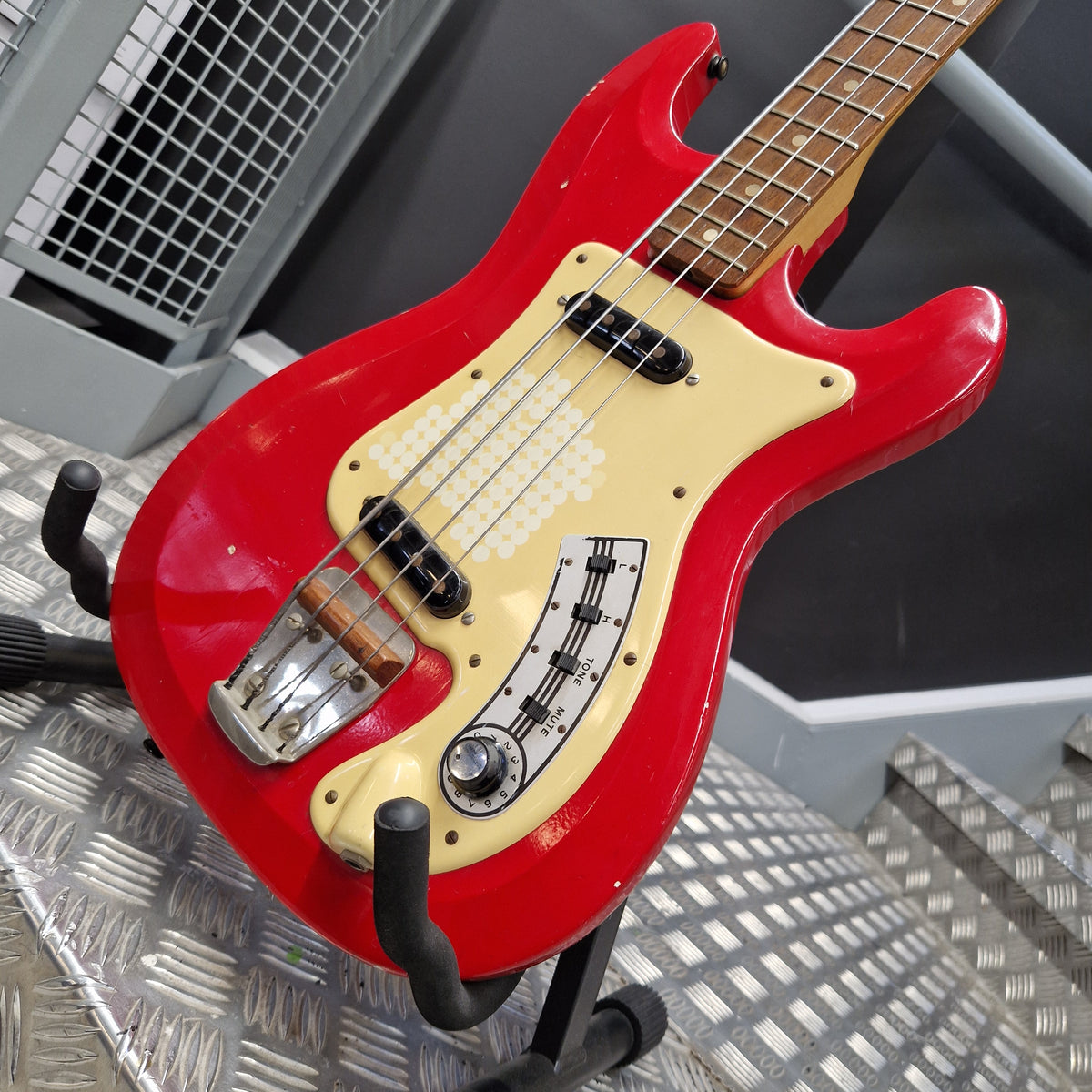 Pre-Owned Hagstrom Futurama Deluxe 1960s Bass - Red A red electric bass guitar with a cream-colored pickguard and black hardware, resting on a stand in a workshop setting.