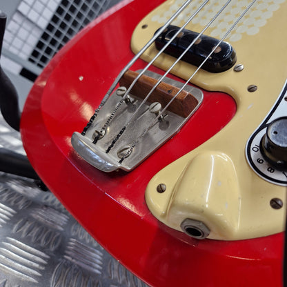 Pre-Owned Hagstrom Futurama Deluxe 1960s Bass - Red Close-up of a red electric guitar body with a cream-colored pickguard, black control knobs, and a chrome bridge, resting on a textured metal surface.