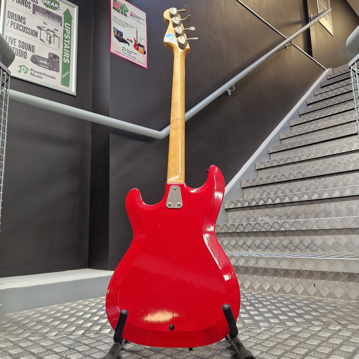 Pre-Owned Hagstrom Futurama Deluxe 1960s Bass - Red A red electric guitar body and light wood neck are shown hanging on a display stand inside a store or showroom with metal stairs and informational signs in the background.
