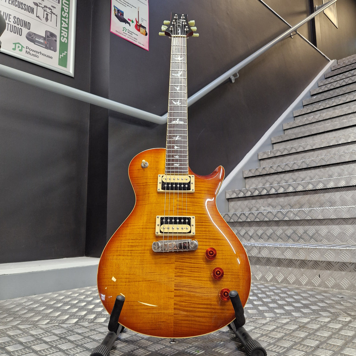 Pre-Owned PRS SE Bernie Marsden Signature Electric - Vintage Sunburst A sunburst electric guitar with gold hardware and a black pickguard, mounted on a display stand in a gallery setting with informational signs and textured walls.