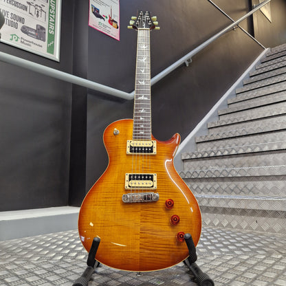 Pre-Owned PRS SE Bernie Marsden Signature Electric - Vintage Sunburst A sunburst electric guitar with gold hardware and a black pickguard, mounted on a display stand in a gallery setting with informational signs and textured walls.