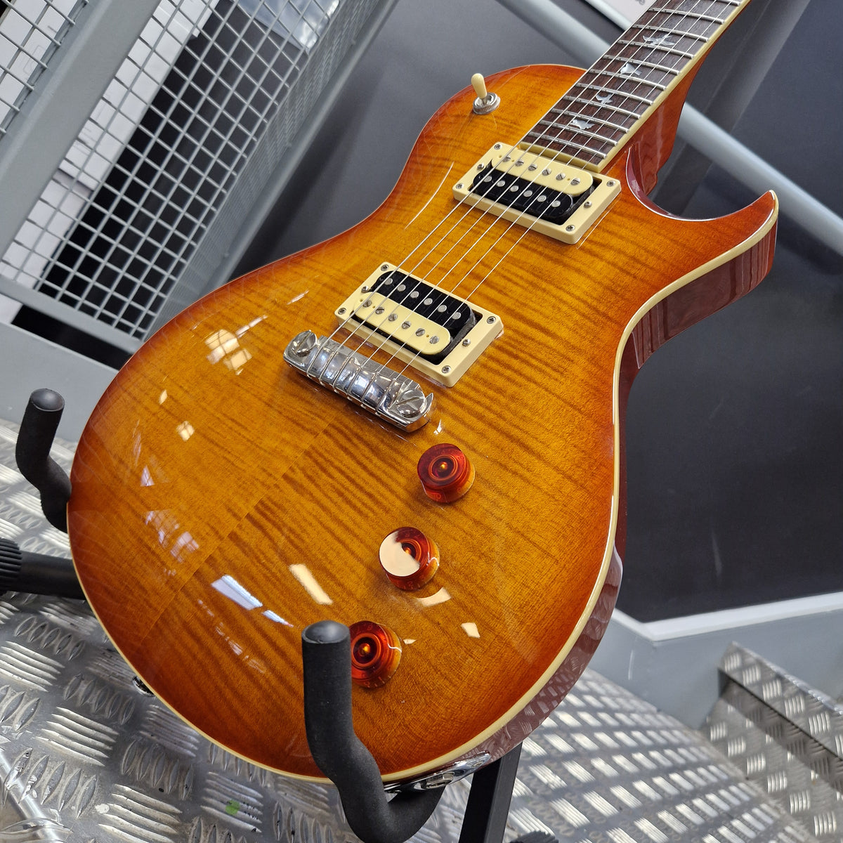Pre-Owned PRS SE Bernie Marsden Signature Electric - Vintage Sunburst A close-up of a PRS electric guitar body in a glossy amber-orange flamed maple finish, resting on a stand. The guitar features gold hardware, red control knobs, and a black pickguard, set against a gray industrial background.