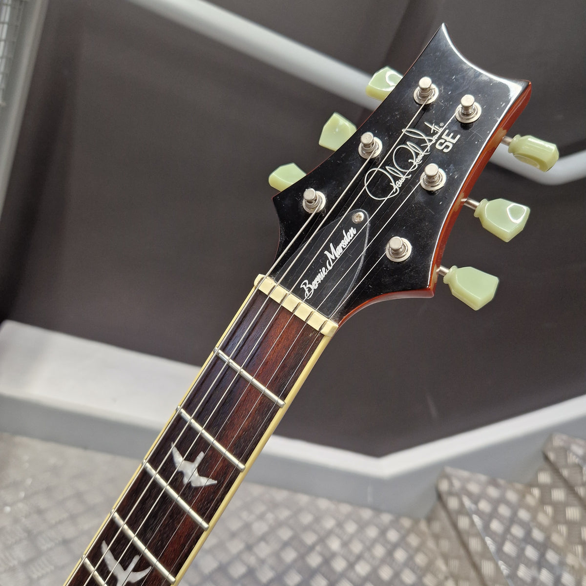 Pre-Owned PRS SE Bernie Marsden Signature Electric - Vintage Sunburst Close-up of a guitar headstock and upper fretboard, showing a black headstock with light green tuning pegs, gold hardware, and white script branding on a dark background.