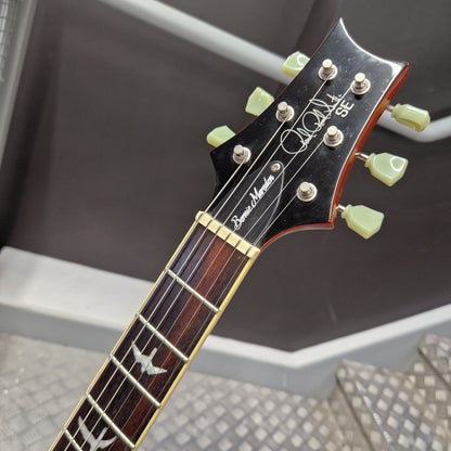 Pre-Owned PRS SE Bernie Marsden Signature Electric - Vintage Sunburst Close-up of a guitar headstock and upper fretboard, showing a black headstock with light green tuning pegs, gold hardware, and white script branding on a dark background.