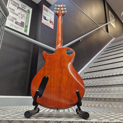 Pre-Owned PRS SE Bernie Marsden Signature Electric - Vintage Sunburst A reddish-brown electric guitar shown from the back, mounted on a black stand inside a store or showroom with metal walls and informational posters visible.