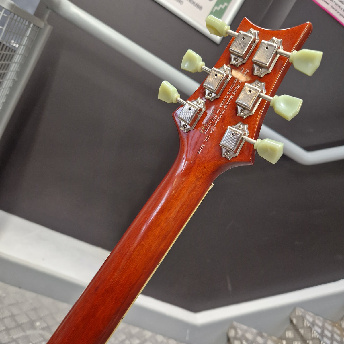 Pre-Owned PRS SE Bernie Marsden Signature Electric - Vintage Sunburst Close-up of the headstock and upper neck of a glossy reddish-brown electric guitar, showing six light green tuning pegs and engraved model text on a dark display surface.