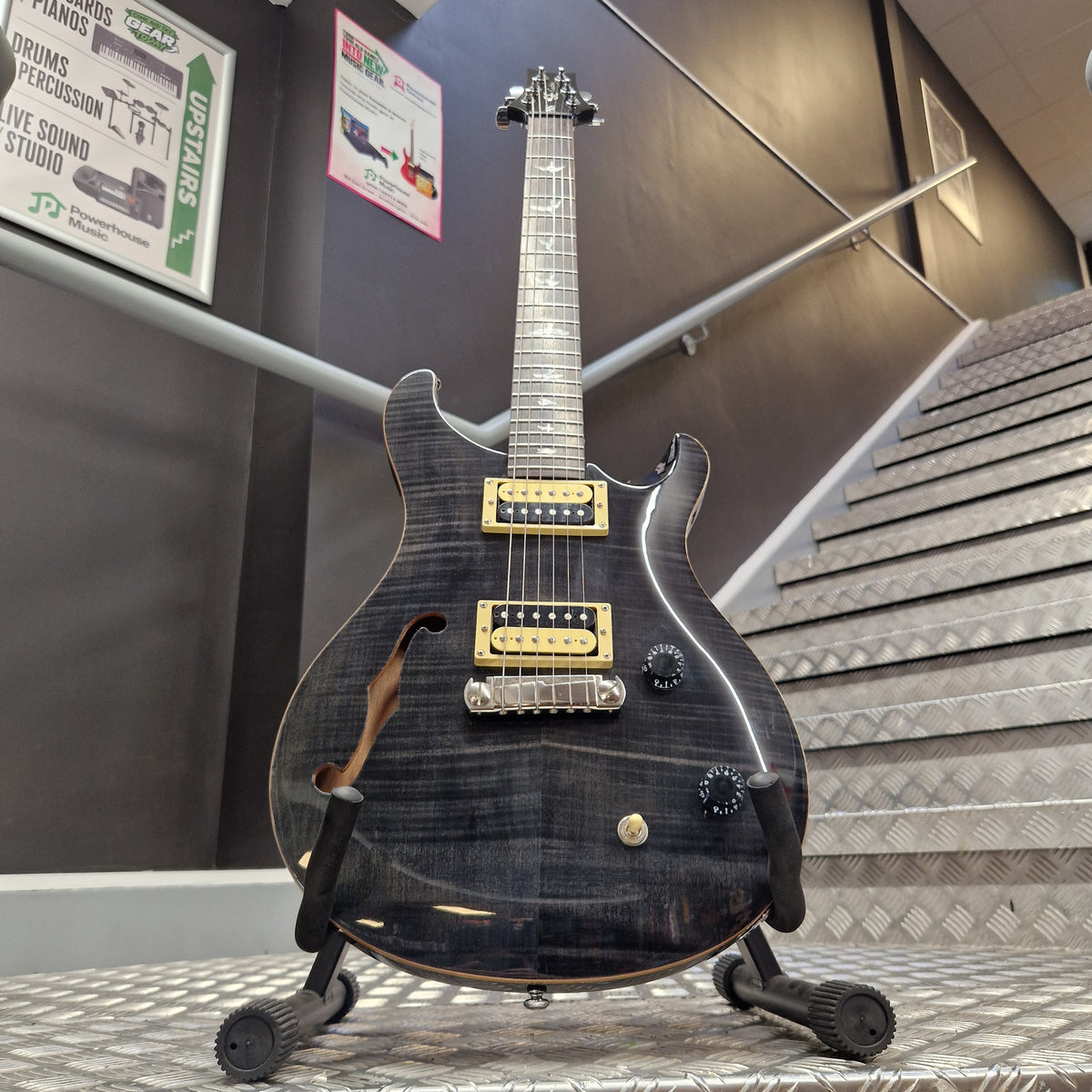 Pre-Owned PRS SE Custom 22 Semi-Hollow Electric Korean - Grey Black A black electric guitar with gold hardware and a flamed maple top, displayed on a stand inside what appears to be a showroom or shop.