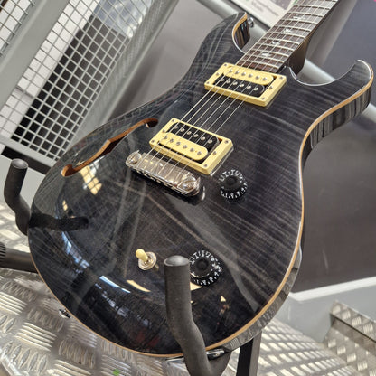 Pre-Owned PRS SE Custom 22 Semi-Hollow Electric Korean - Grey Black A close-up of a black electric guitar with a glossy flamed maple top, gold hardware, and black controls, resting on a stand.