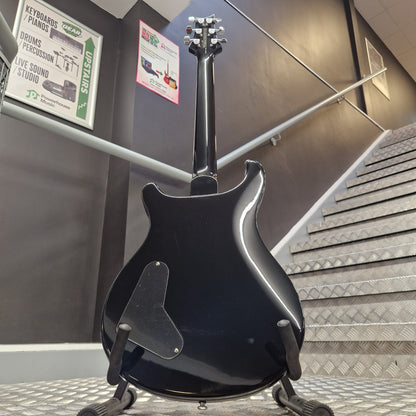 Pre-Owned PRS SE Custom 22 Semi-Hollow Electric Korean - Grey Black A glossy black electric guitar body on a display stand in an indoor showroom or warehouse setting with metal stairs and informational signs visible in the background.