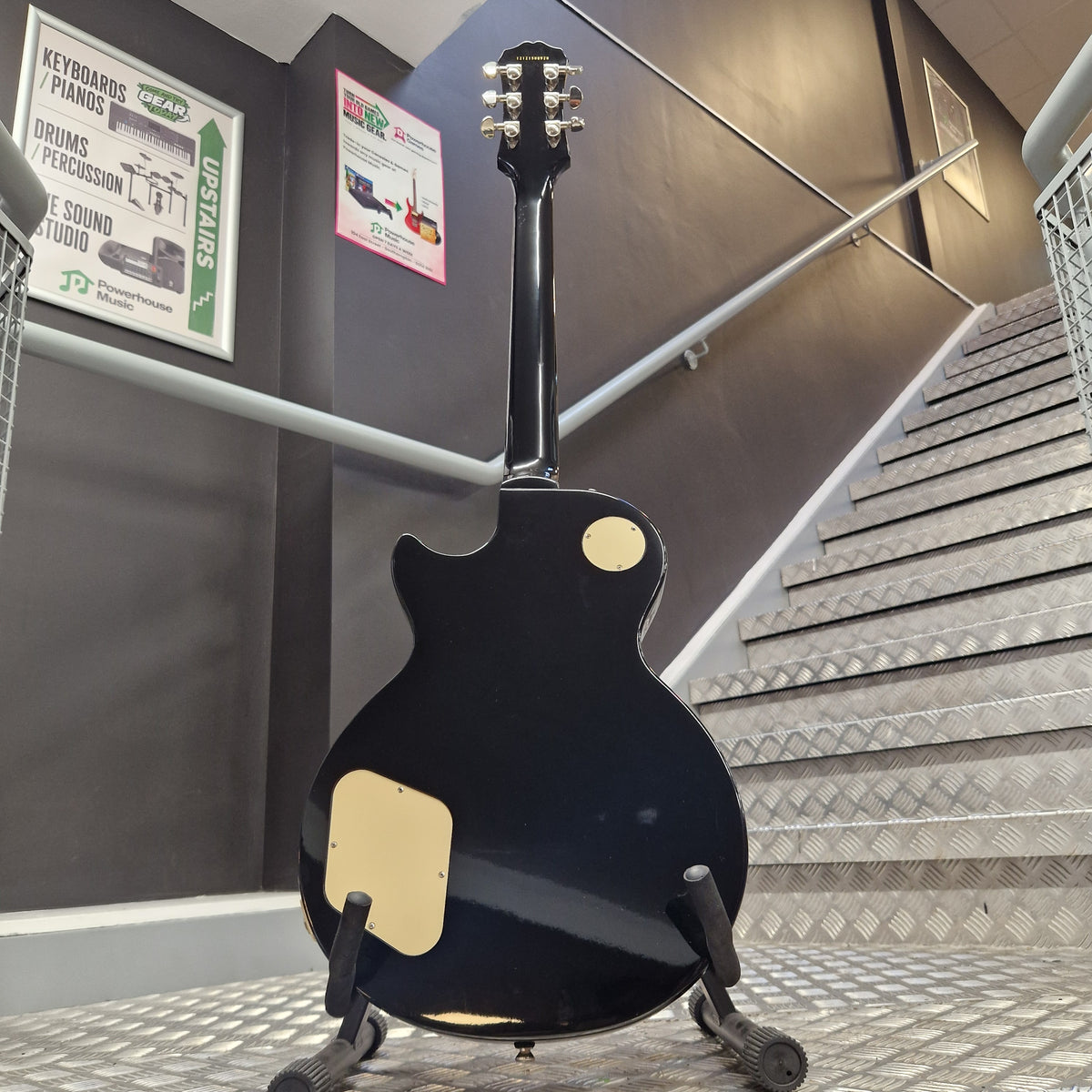 Pre-Owned Epiphone Les Paul Standard Pro Electric - Vintage Sunburst A black electric guitar body and neck are shown from the back, mounted on a stand in an indoor industrial setting with metal stairs and informational posters visible in the background.