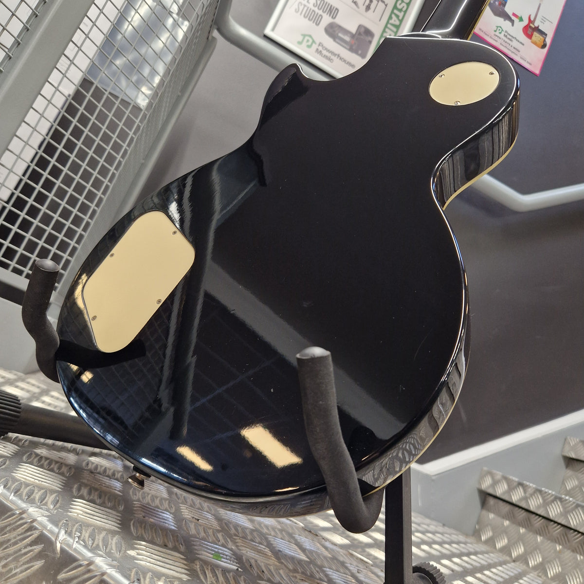 Pre-Owned Epiphone Les Paul Standard Pro Electric - Vintage Sunburst A glossy black electric guitar body viewed from the back, resting on a stand in a store or workshop setting with metal diamond plate flooring visible.