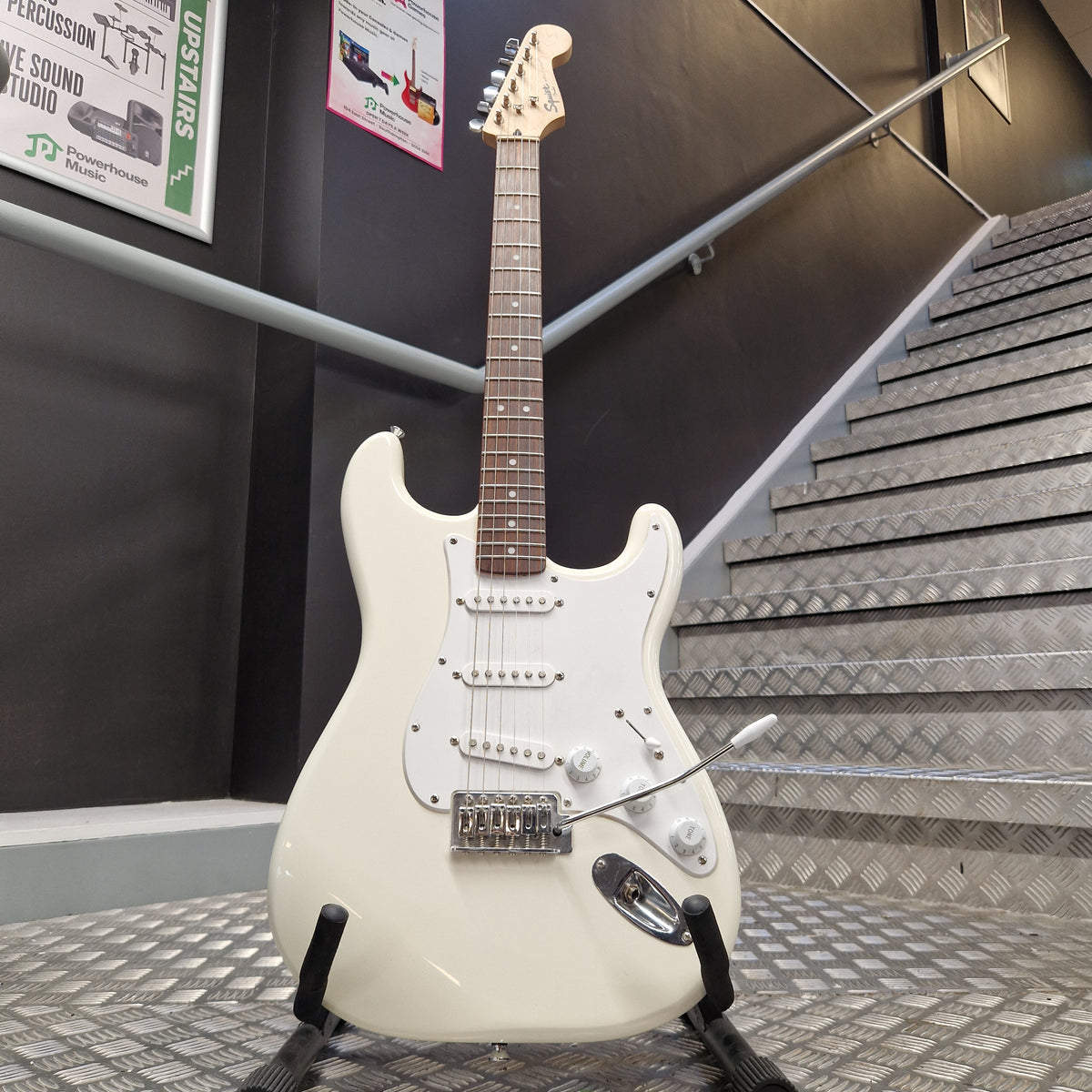 Pre-Owned Squier Bullet Stratocaster Electric - Arctic White A white electric guitar with a dark brown fretboard and black hardware, hanging on a wall in an indoor setting with metal paneling.