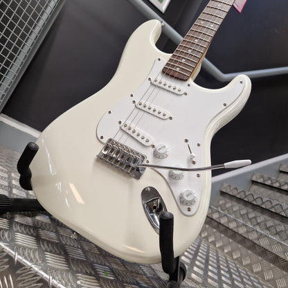 Pre-Owned Squier Bullet Stratocaster Electric - Arctic White An off-white electric guitar with chrome hardware and a black pickguard, resting on a stand in a shop setting with a diamond plate wall in the background.
