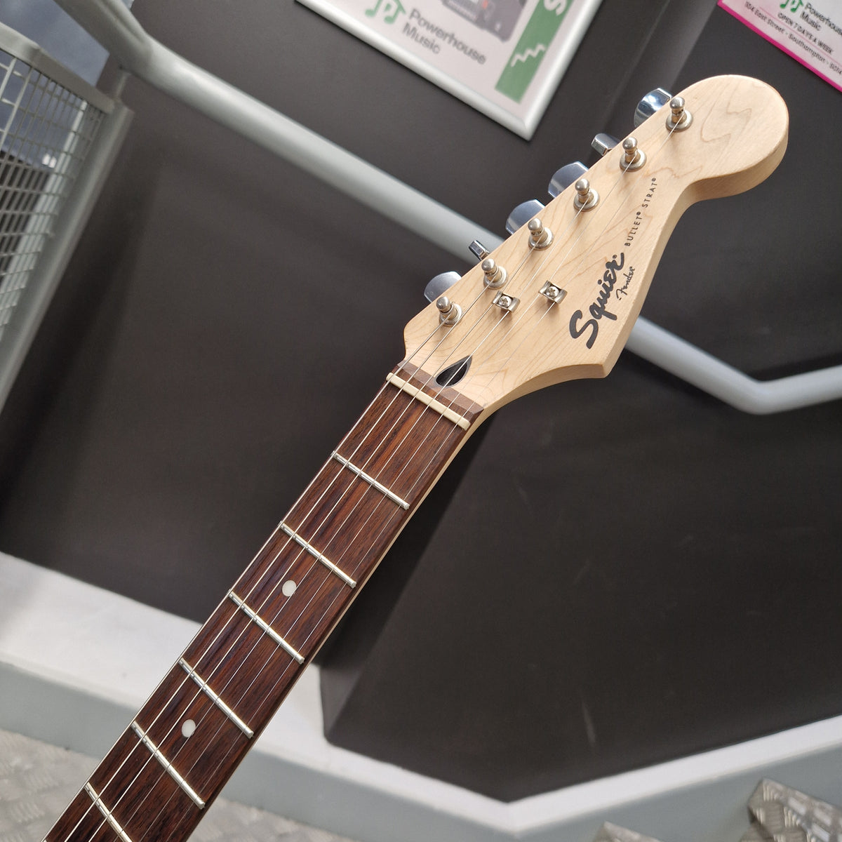 Pre-Owned Squier Bullet Stratocaster Electric - Arctic White Close-up of a Squier electric guitar headstock and fretboard, showing the natural wood headstock with six silver tuning pegs and the Squier logo, resting on a dark display stand.