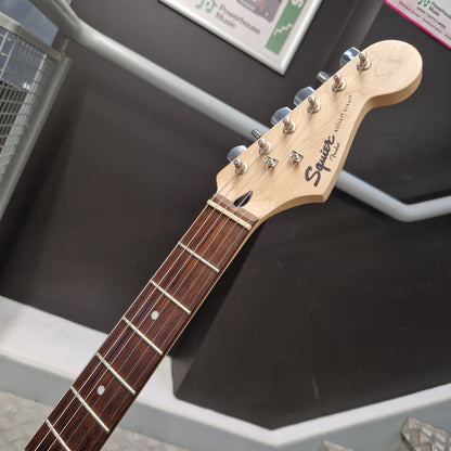 Pre-Owned Squier Bullet Stratocaster Electric - Arctic White Close-up of a Squier electric guitar headstock and fretboard, showing the natural wood headstock with six silver tuning pegs and the Squier logo, resting on a dark display stand.