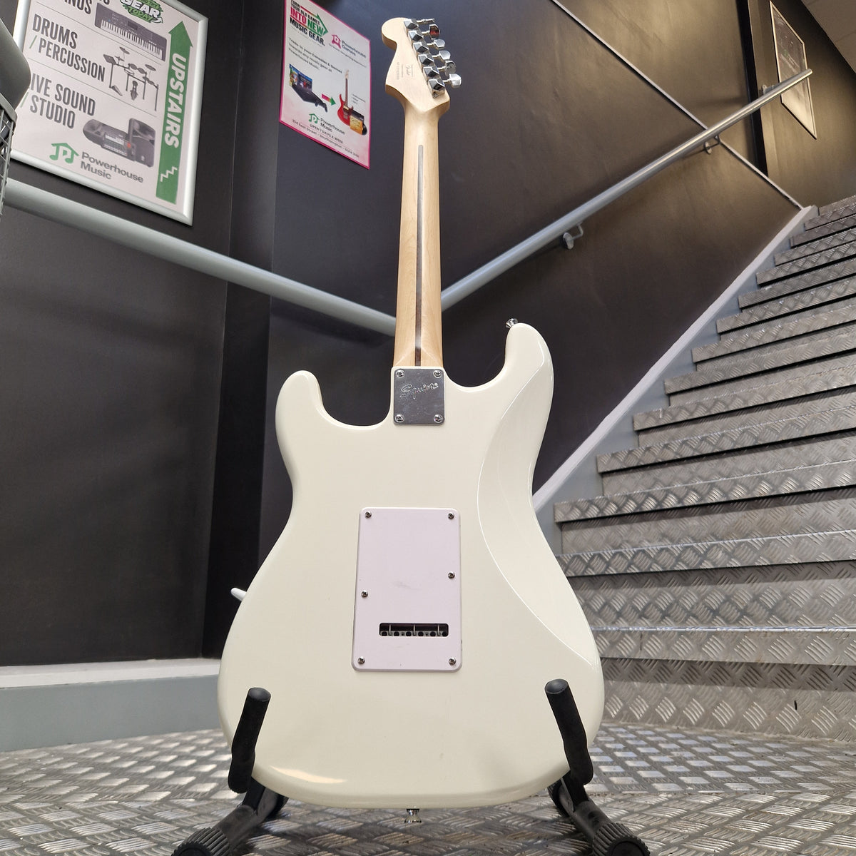 Pre-Owned Squier Bullet Stratocaster Electric - Arctic White An electric guitar body in off-white/cream finish, photographed from above on a display stand inside what appears to be a showroom or studio. The neck is light wood, and the back has a white pickup cover.