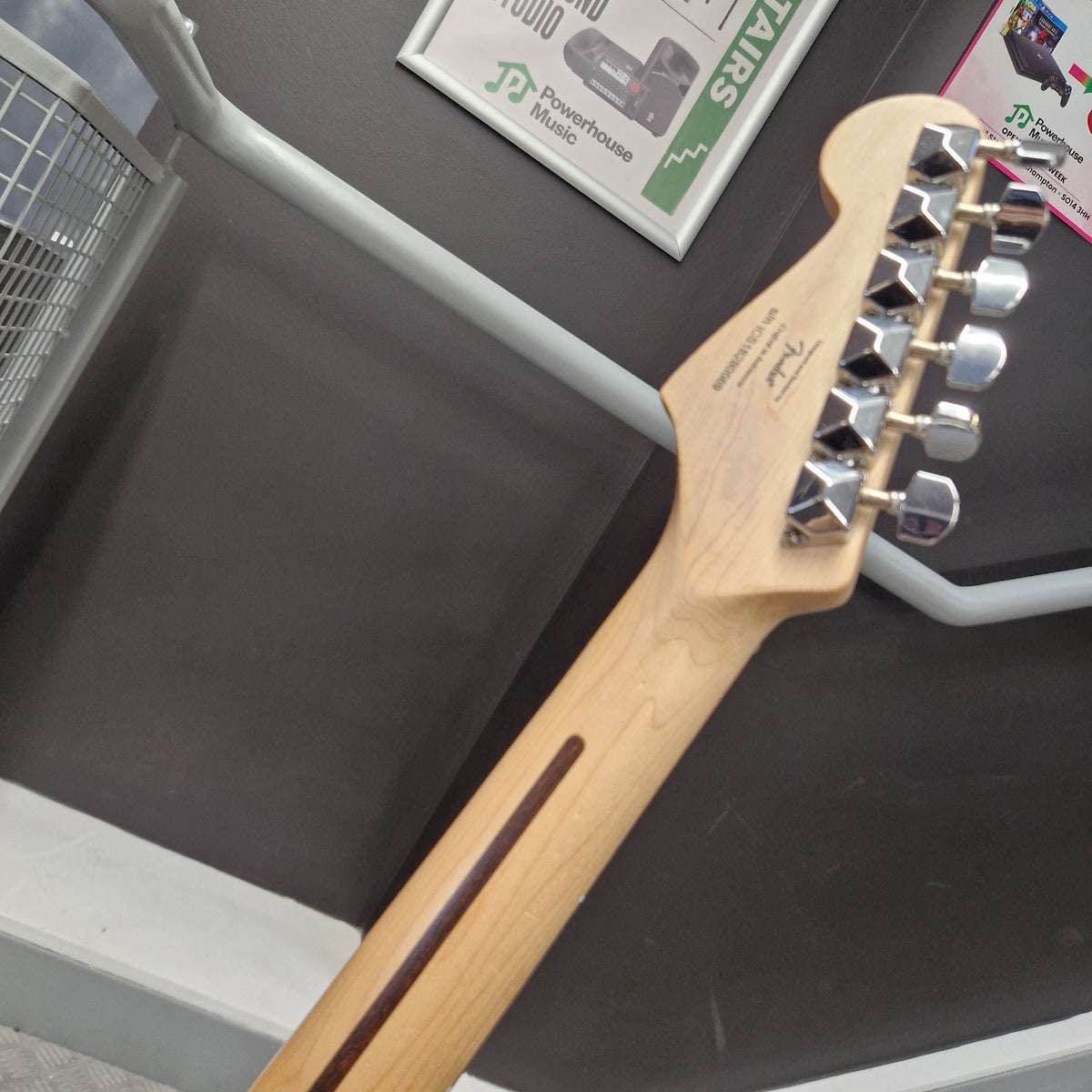 Pre-Owned Squier Bullet Stratocaster Electric - Arctic White Close-up of a Fender electric guitar headstock and neck, showing a natural light wood finish, chrome tuning pegs, and a dark brown inlay on a dark gray surface.