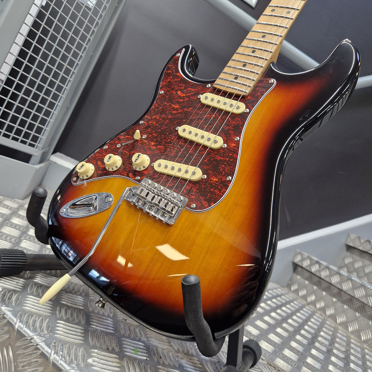 Pre-Owned Jet JS-300 Left-Handed Electric - Sunburst A Fender electric guitar with a classic sunburst finish, resting on a stand in front of a textured metal background.