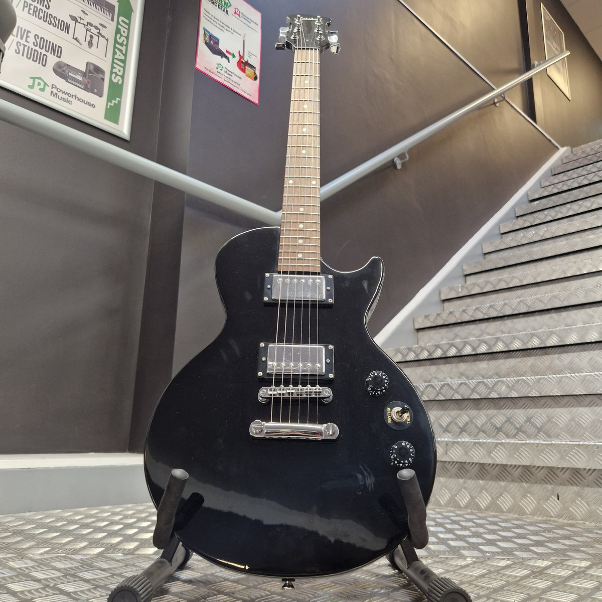 Pre-Owned Marshall MGAP Singlecut Electric - Black Gloss A black electric guitar with chrome hardware and a light wood fretboard, displayed on a stand in an indoor setting with metal paneling and informational signs in the background.