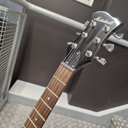Pre-Owned Marshall MGAP Singlecut Electric - Black Gloss Close-up of a Marshall electric guitar headstock and fretboard, showing the brand name in script and six silver tuning pegs against a dark indoor background.
