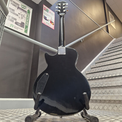 Pre-Owned Marshall MGAP Singlecut Electric - Black Gloss A black electric guitar shown from the back, mounted on a display stand in an indoor showroom or warehouse setting with metal stairs and informational signs visible in the background.