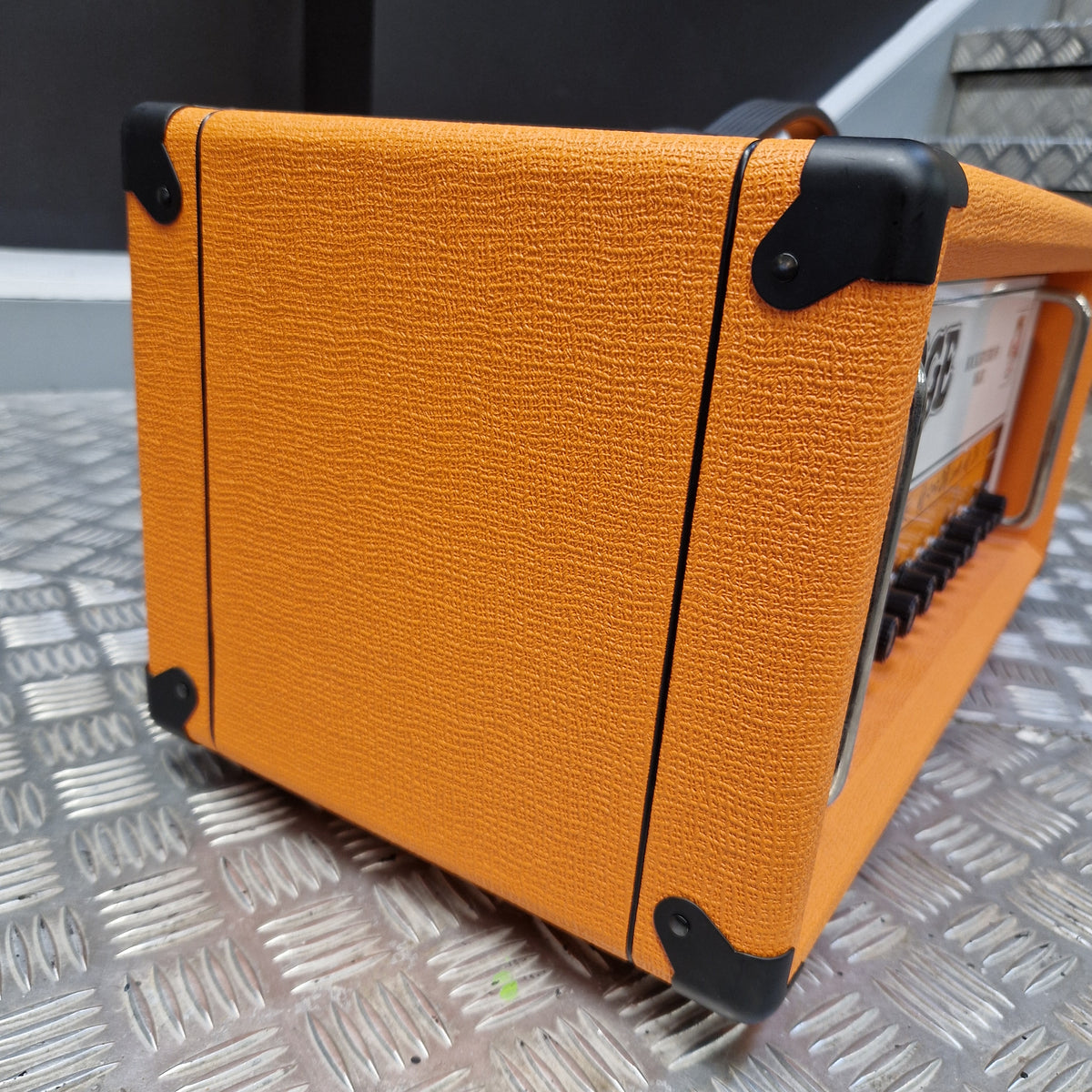 Pre-Owned Orange Rockerverb 50H MKIII Valve Electric Amplifier Head An orange textured guitar amplifier or speaker cabinet with black corner protectors and a metal handle, photographed on a diamond-plate metal surface.