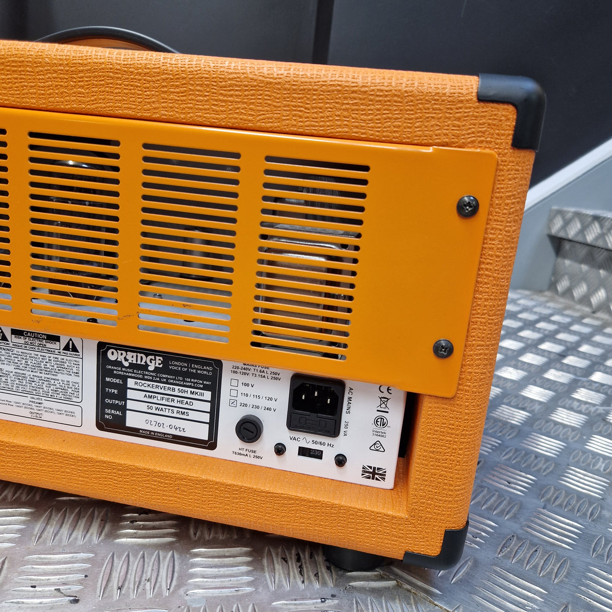 Pre-Owned Orange Rockerverb 50H MKIII Valve Electric Amplifier Head Close-up of the back of an Orange guitar amplifier, showing the orange textured cabinet, vent grille, power input, and control labels, resting on a diamond plate metal surface.