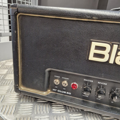 Pre-Owned Blackstar HT Club 50 Electric All-Valve Amplifier Head Close-up of a black vintage-style guitar amplifier head with gold trim, black knobs, a red indicator light, and the partial visible 'Blas' logo, resting on a diamond plate metal surface.