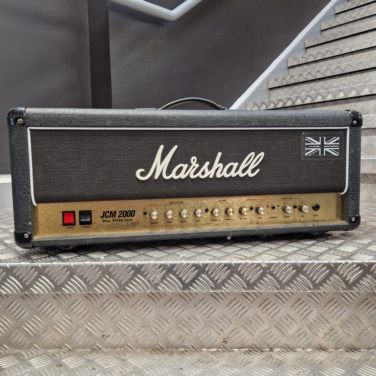 Pre-Owned Marshall JCM2000 DSL100 Valve Electric Amplifier Head A Marshall JCM 2000 guitar amplifier head photographed from above on a diamond-plate metal surface. The head has a black front grille with the white Marshall logo and Union Jack emblem, plus a tan control panel with knobs and a red switch.