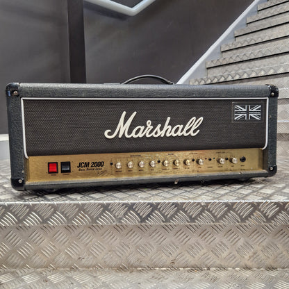 Pre-Owned Marshall JCM2000 DSL100 Valve Electric Amplifier Head A Marshall JCM 2000 guitar amplifier head photographed from above on a diamond-plate metal surface. The head has a black front grille with the white Marshall logo and Union Jack emblem, plus a tan control panel with knobs and a red switch.