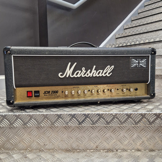 Pre-Owned Marshall JCM2000 DSL100 Valve Electric Amplifier Head A Marshall JCM 2000 guitar amplifier head photographed from above on a diamond-plate metal surface. The head has a black front grille with the white Marshall logo and Union Jack emblem, plus a tan control panel with knobs and a red switch.