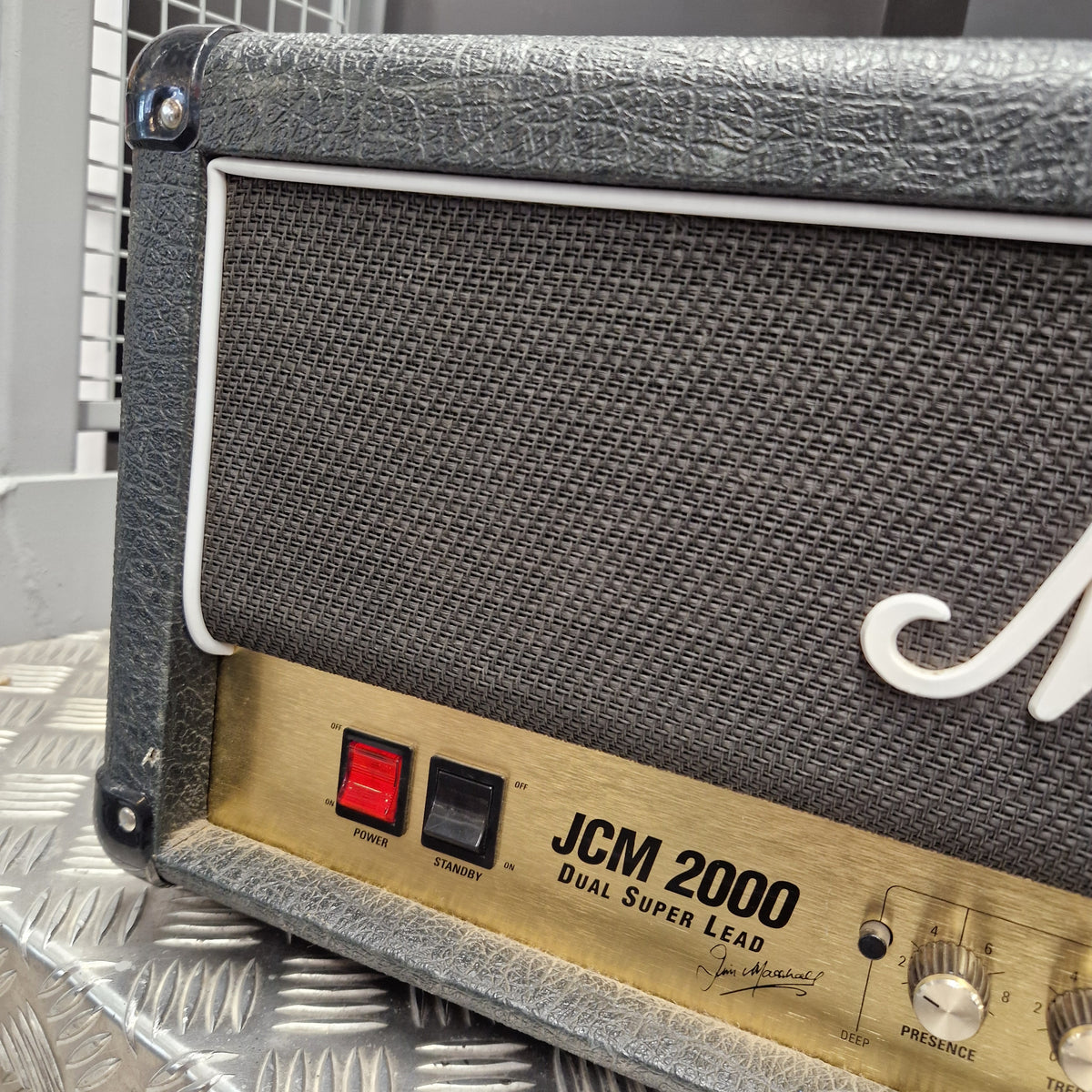 Pre-Owned Marshall JCM2000 DSL100 Valve Electric Amplifier Head Close-up of a guitar amplifier front panel with a black textured grille, white trim, and a gold control panel labeled JCM 2000 Dual Super Lead.