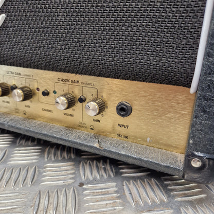 Pre-Owned Marshall JCM2000 DSL100 Valve Electric Amplifier Head Close-up of a vintage guitar amplifier control panel with a brushed gold metal faceplate, black mesh grille, and silver knobs, resting on a diamond plate metal surface.