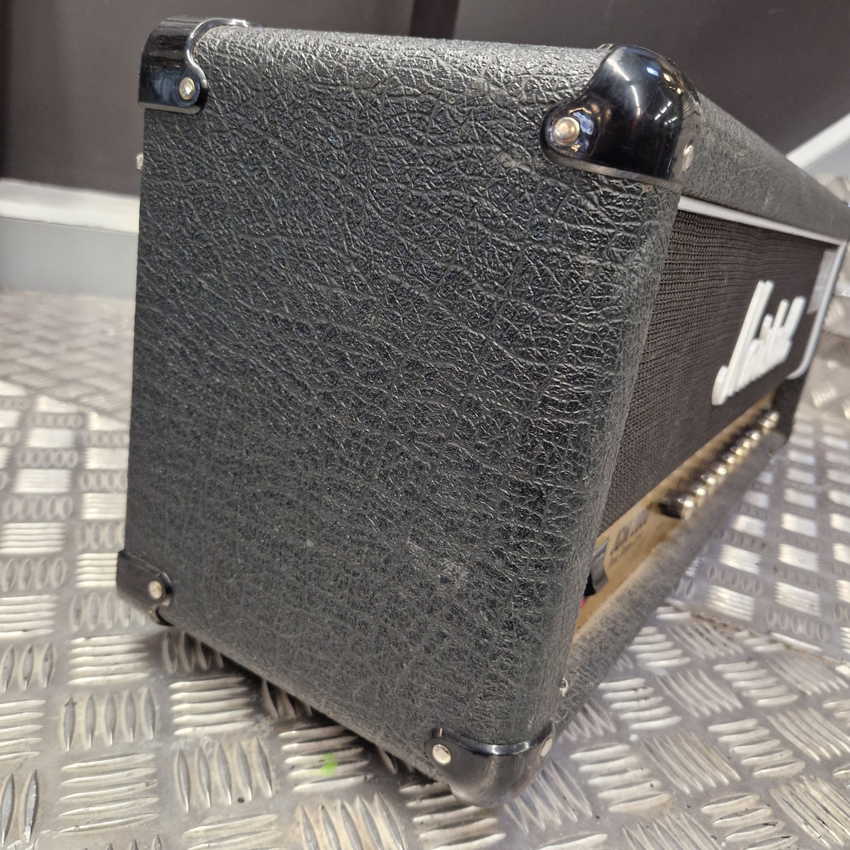 Pre-Owned Marshall JCM2000 DSL100 Valve Electric Amplifier Head A close-up shot of a black textured guitar amplifier head, showing the front grille, corner protectors, and part of the gold-toned metal side frame, resting on a silver diamond plate metal surface.