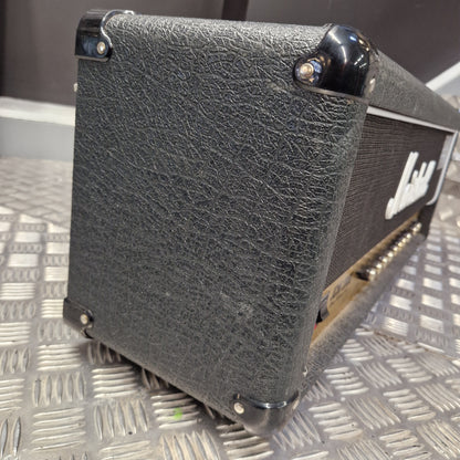 Pre-Owned Marshall JCM2000 DSL100 Valve Electric Amplifier Head A close-up shot of a black textured guitar amplifier head, showing the front grille, corner protectors, and part of the gold-toned metal side frame, resting on a silver diamond plate metal surface.